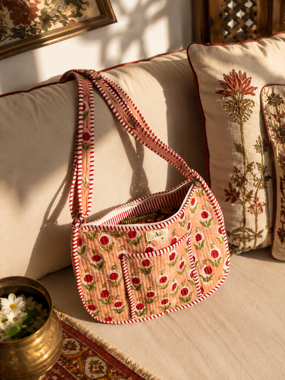 A peach-coloured quilted crossbody shoulder bag with a red and white striped border and a floral print, resting on a beige sofa with decorative pillows in a warm, sunlit room.