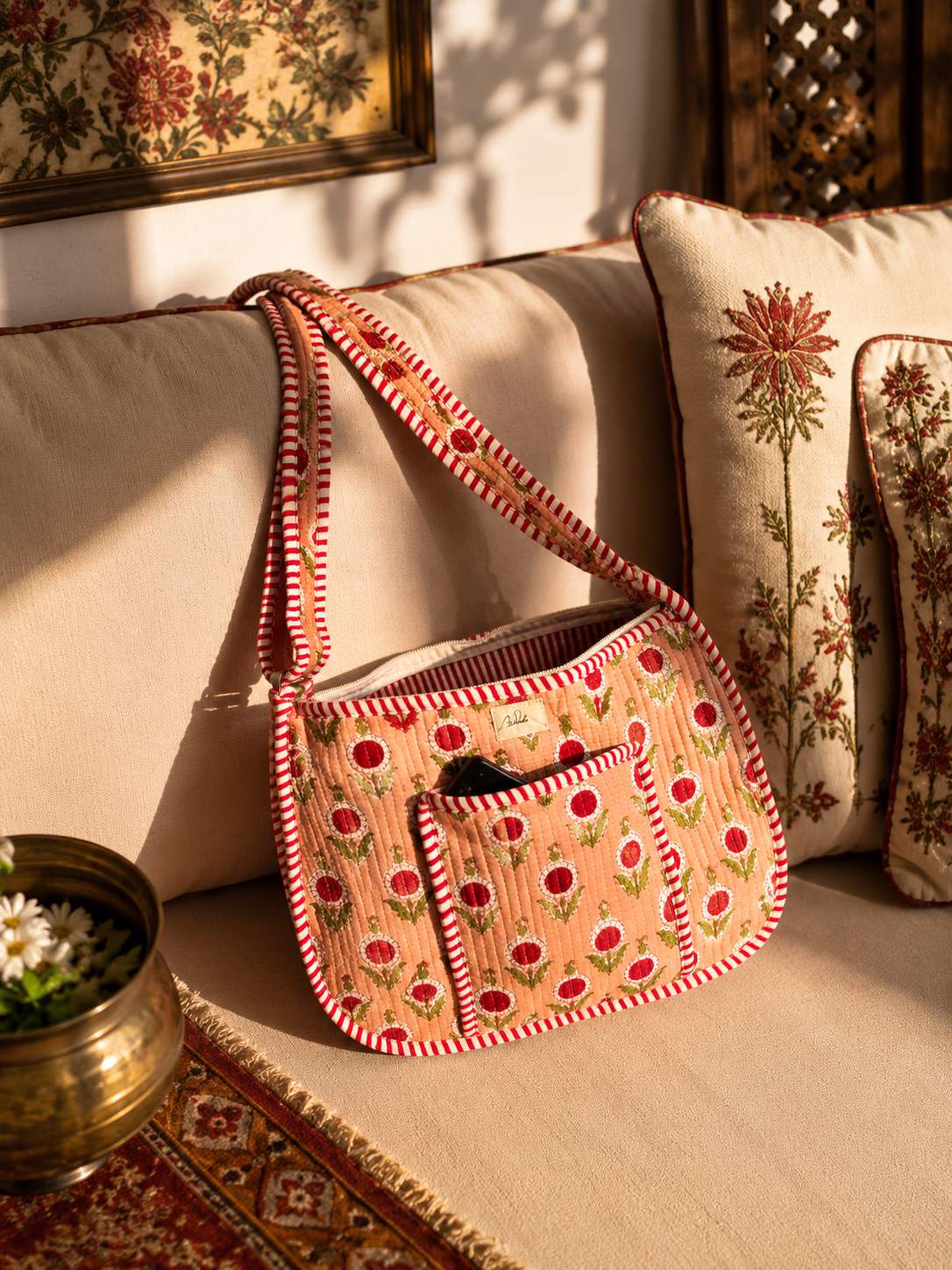 A quilted peach crossbody shoulder bag with red and white striped trim and red floral motifs, with a phone in the front pocket and open chain, resting on a beige sofa with decorative pillows in a warm, sunlit room.