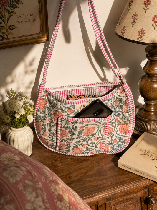 A white crescent-shaped crossbody bag with a lotus and leaves all-over pattern with stripped interiors and trims with a book inside and a mobile in the front pocket, resting on a wooden table with a wooden table lamp and flower vase. 