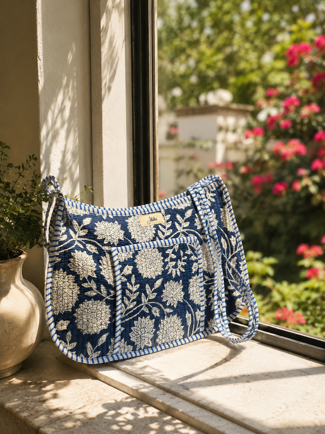 A blue and white floral quilted crossbody shoulder bag with a braided strap, resting on a stone windowsill with a garden view in the background.