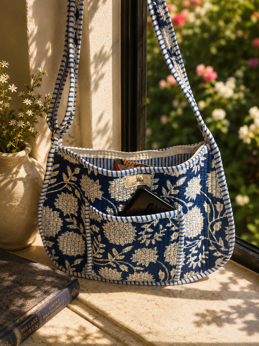 A navy blue and white floral patterned shoulder bag with a front pocket holding a phone, styled on a stone windowsill with a potted plant and book nearby.