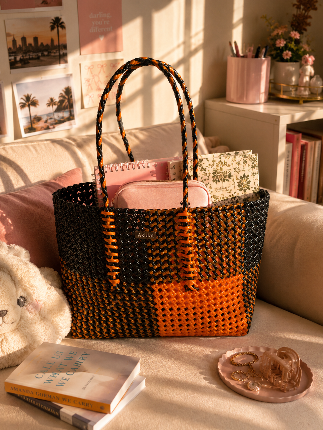 A woven recycled plastic tote bag with black and orange checkered pattern and double shoulder straps, sitting on a beige sofa in a warm, sunlit room with books, a teddy bear, and stationery around it.