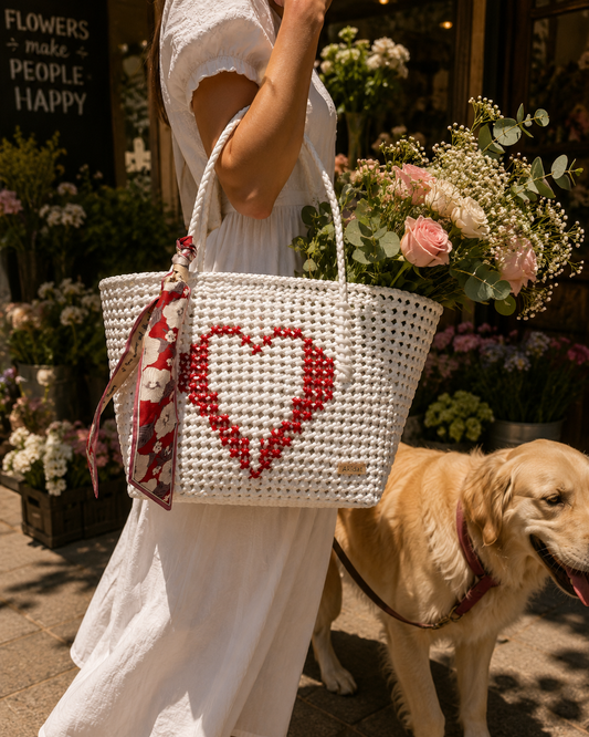 A woman in a white dress carries a white recycled plastic woven tote bag with a red heart design and a red patterned scarf. A golden retriever stands beside her holding flowers in front of a flower shop.