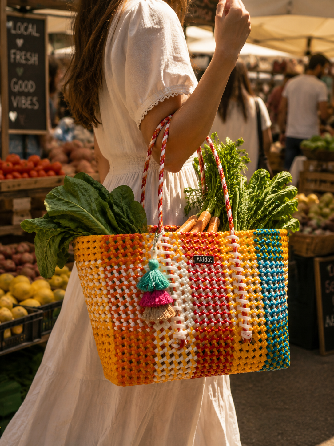 A recycled plastic tote bag in the colours orange, red, blue, yellow and white carrying vegetables being carried by a woman wearing a white dress in a farmers' market. 