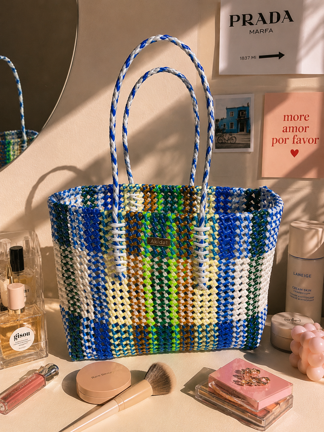 A woven recycled plastic tote bag with blue and white braided handles, featuring a multicolour plaid pattern of blue, green, yellow, and white. The bag is styled on a tabletop with cosmetics, perfume, and a mirror in a warm, sunlit room.