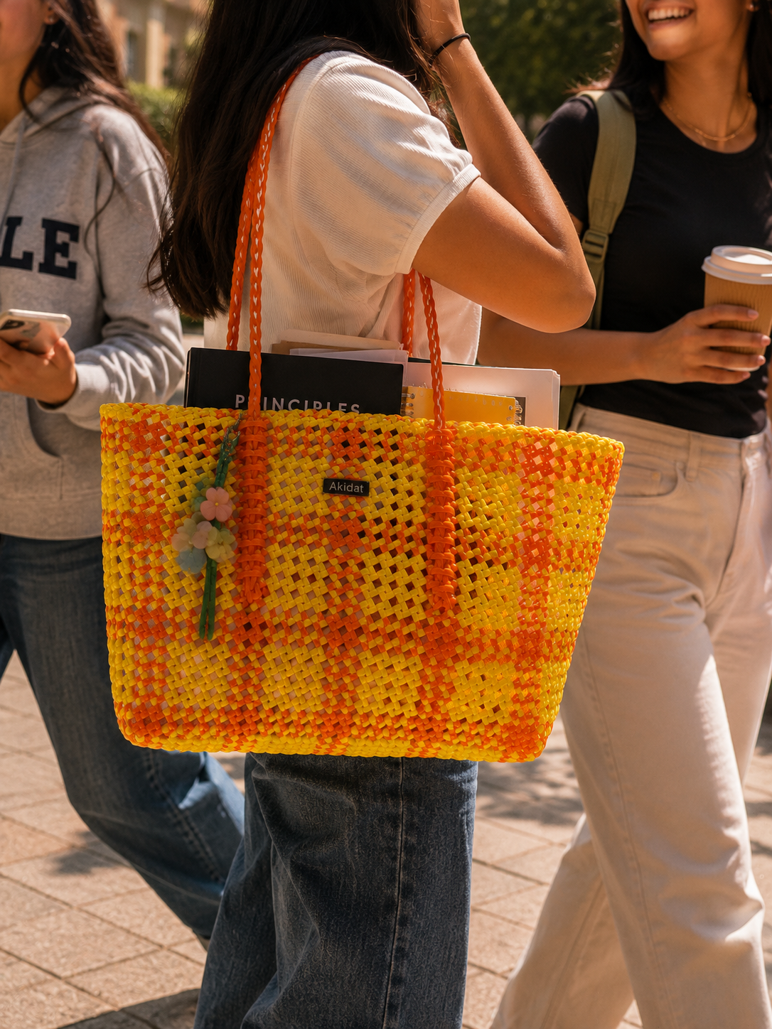 A bright yellow and orange woven recycled plastic tote bag with a plaid pattern, carried on the shoulder of a woman walking outdoors. The bag has a small golden metallic label with the brand name 'Akidat' visible on it.