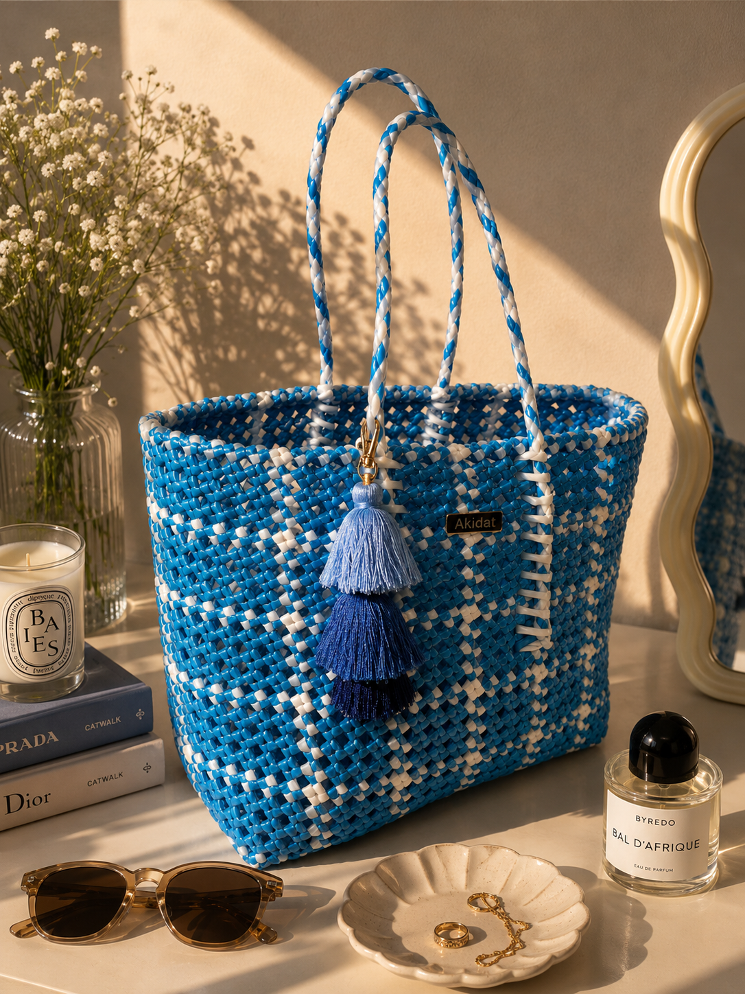 A blue and white recycled plastic woven tote bag with braided handles and two tassels, styled on a tabletop with books, sunglasses, a candle, a perfume bottle, and a mirror in warm sunlight.