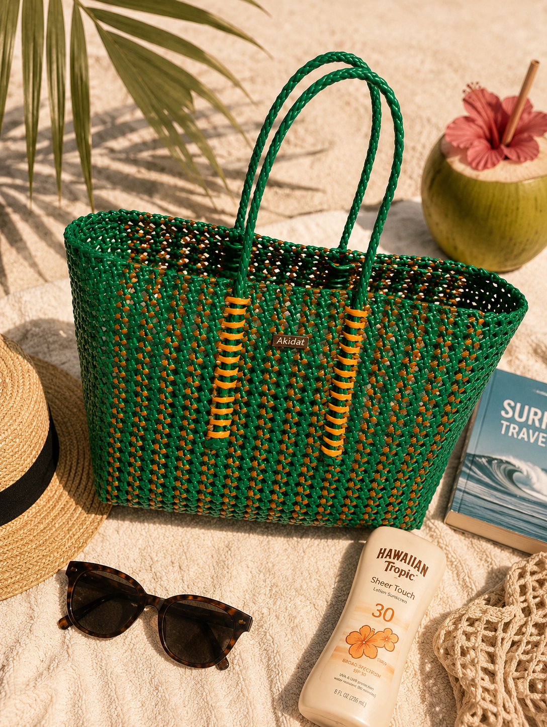 A green woven koodai bag with orange accents made of recycled plastic on a beach surface with sunglasses, sunscreen, and a hat.