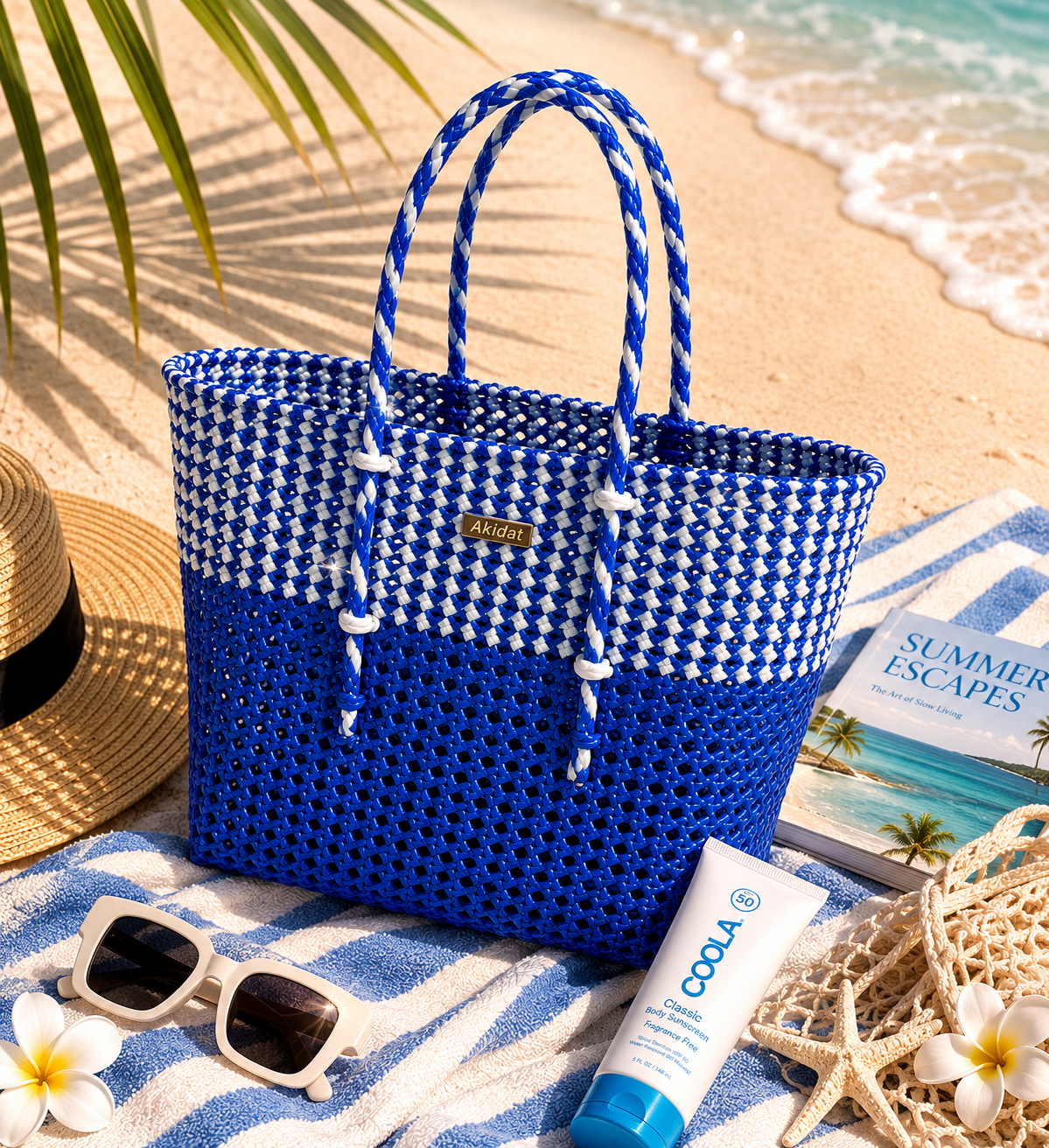 A blue and white woven tote bag made of recycled plastic on a beach with sunglasses, sunscreen, and a book.