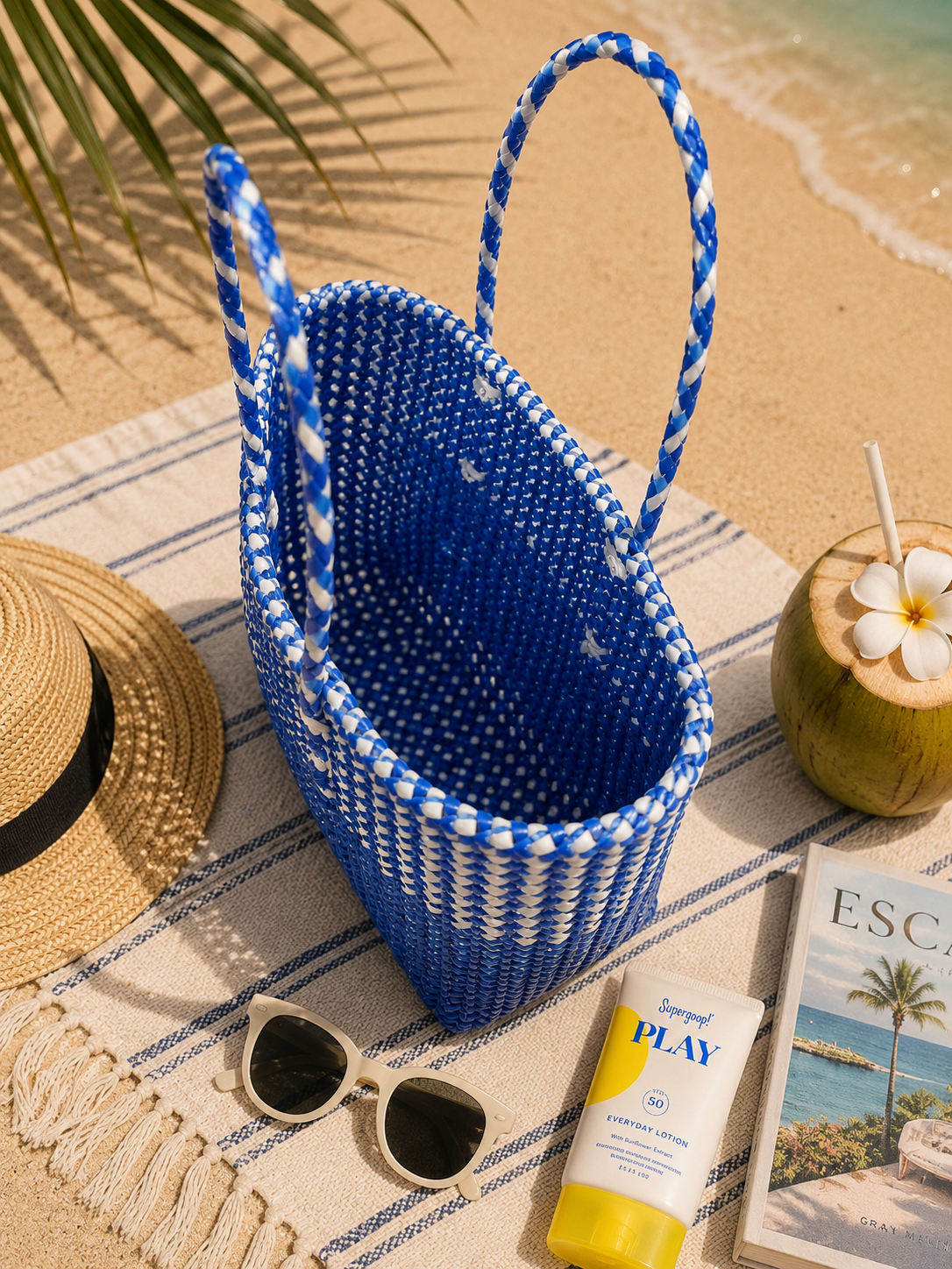 The top view of a blue woven recycled plastic all-weather basket tote on a beach towel with sunglasses, sunscreen, and a magazine.
