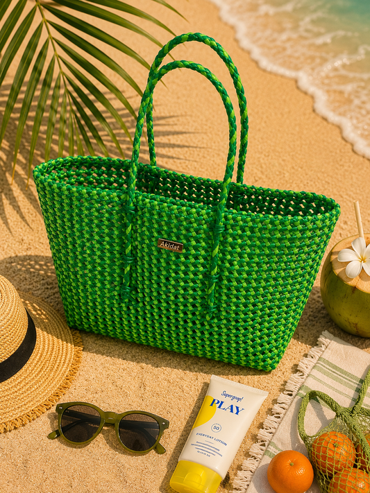 A green and fluorescent green recycled plastic woven tote bag on a beach with sunglasses, sunscreen, and fruit.