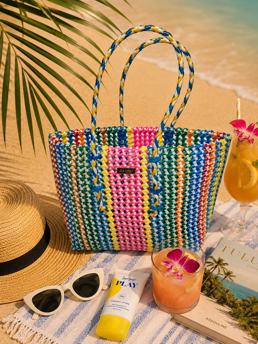 A multicoloured, colourful woven recycled plastic tote bag on a beach with a hat, sunglasses, sunscreen, and a drink.