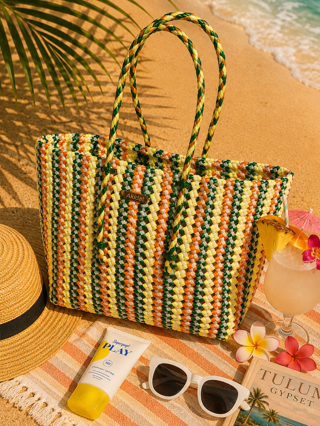 A yellow, orange and green stripe pattern woven with white recycled plastic creates a beautiful tote bag kept on a beach with sunglasses, sunscreen, and a drink.