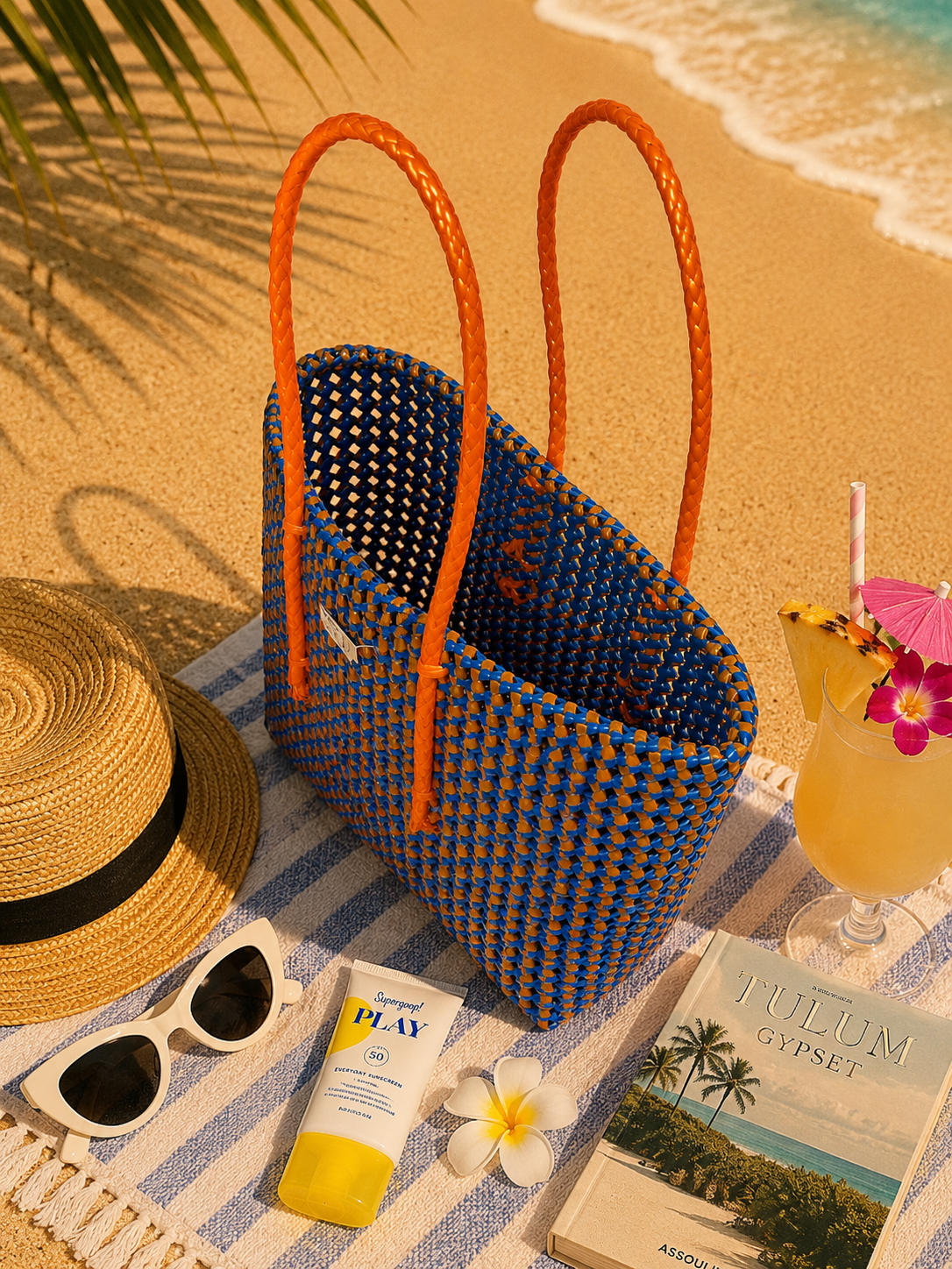 A recycled plastic woven blue and orange bag with orange handles on a striped towel by the beach, with a hat, sunglasses, sunscreen, and a cocktail on it.