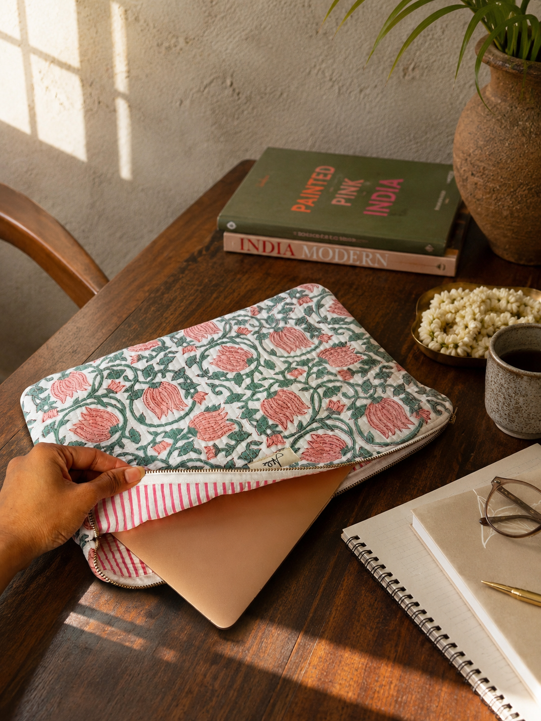 Quilted laptop sleeve with a white base and a pink and green lotus-like blockprint pattern all over with a laptop inside and an internal pink and white stripey pattern, on a wooden study table with a notebook, photo frame, pen holder and some wooden blocks and a mug.