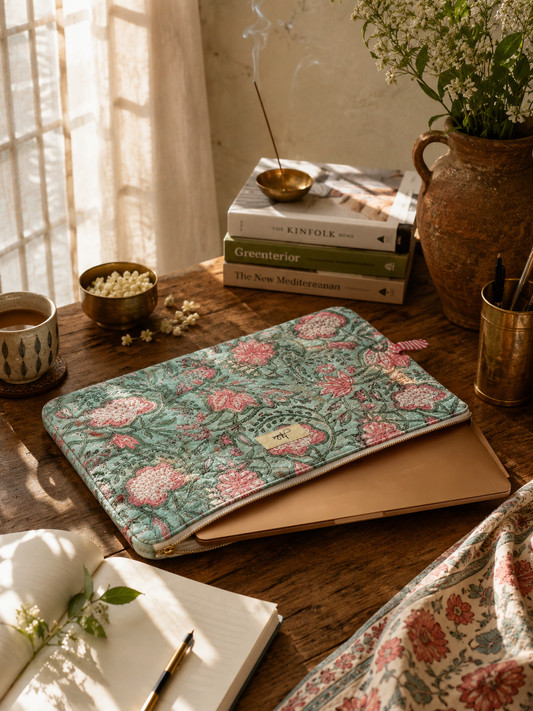 Quilted laptop sleeve with an aqua-coloured base and a pink and green floral blockprint pattern all over with a laptop inside, on a wooden study table with an open notebook, stack of books, pen holder, some wooden blocks and a cup of tea. 