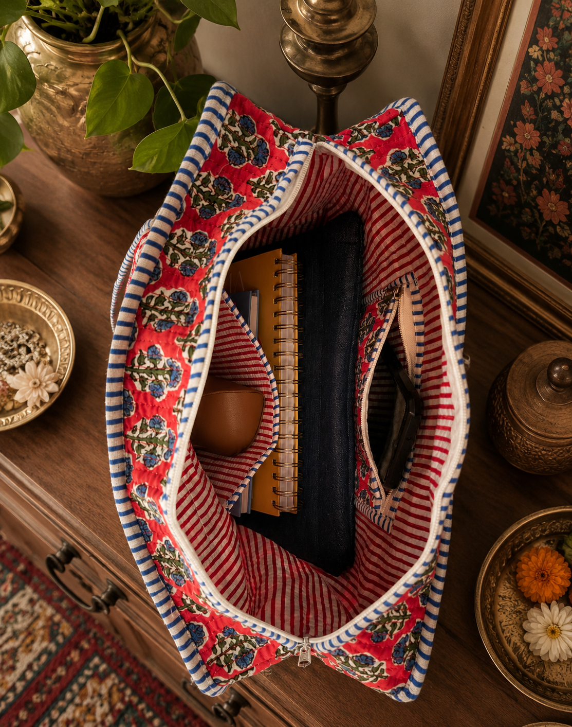 A floral quilted decorative fabric tote bag with a top view with an opened zipper on a wooden surface. The tote bag carries a laptop sleeve, 2 notepads, a sunglass case, and a mobile. 
