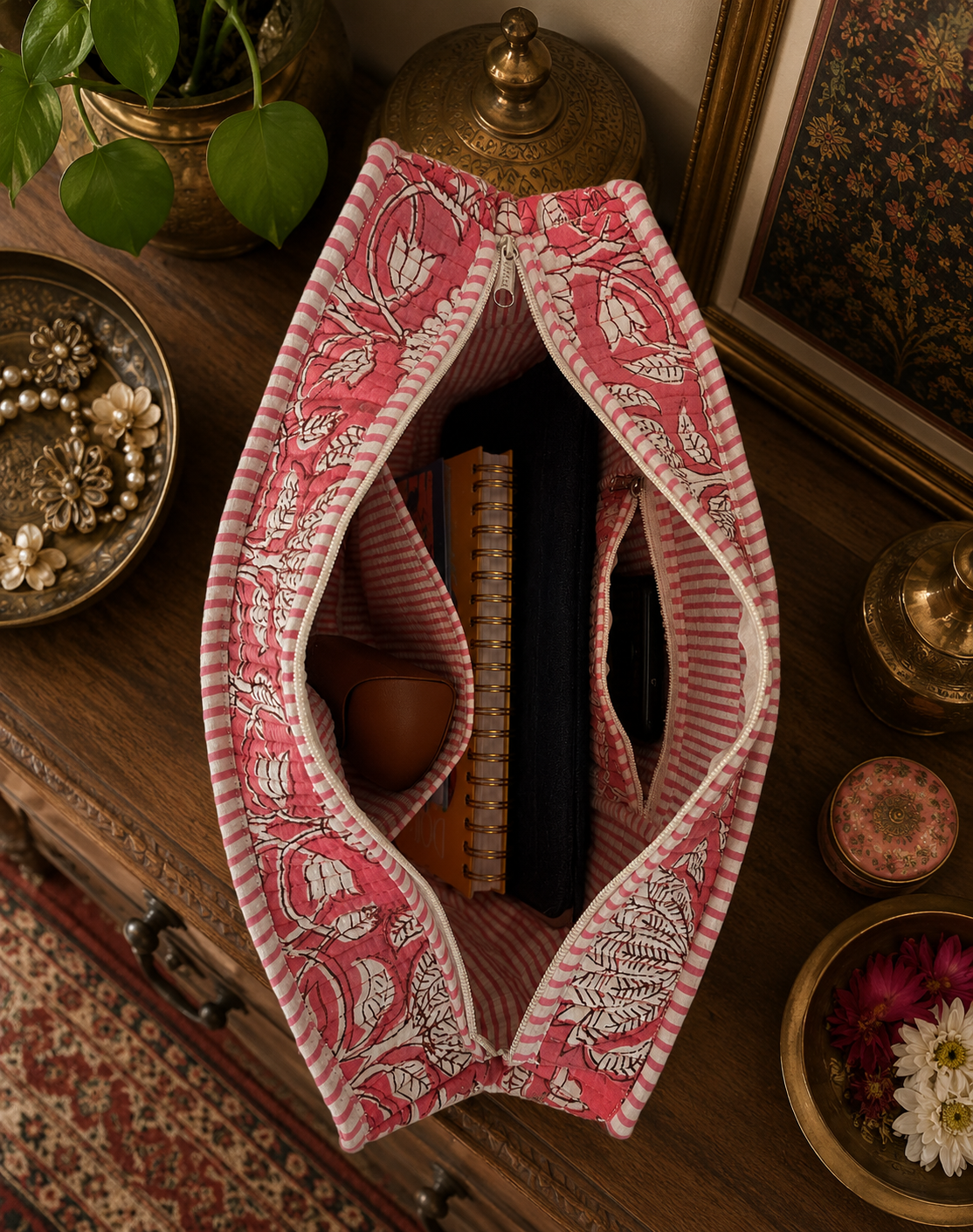 A pink and white patterned block-printed tote bag with an open zipper placed on a wooden surface with decorative items. The bag consists of laptop sleeve, notepads, sunglass cover and a mobile phone. 