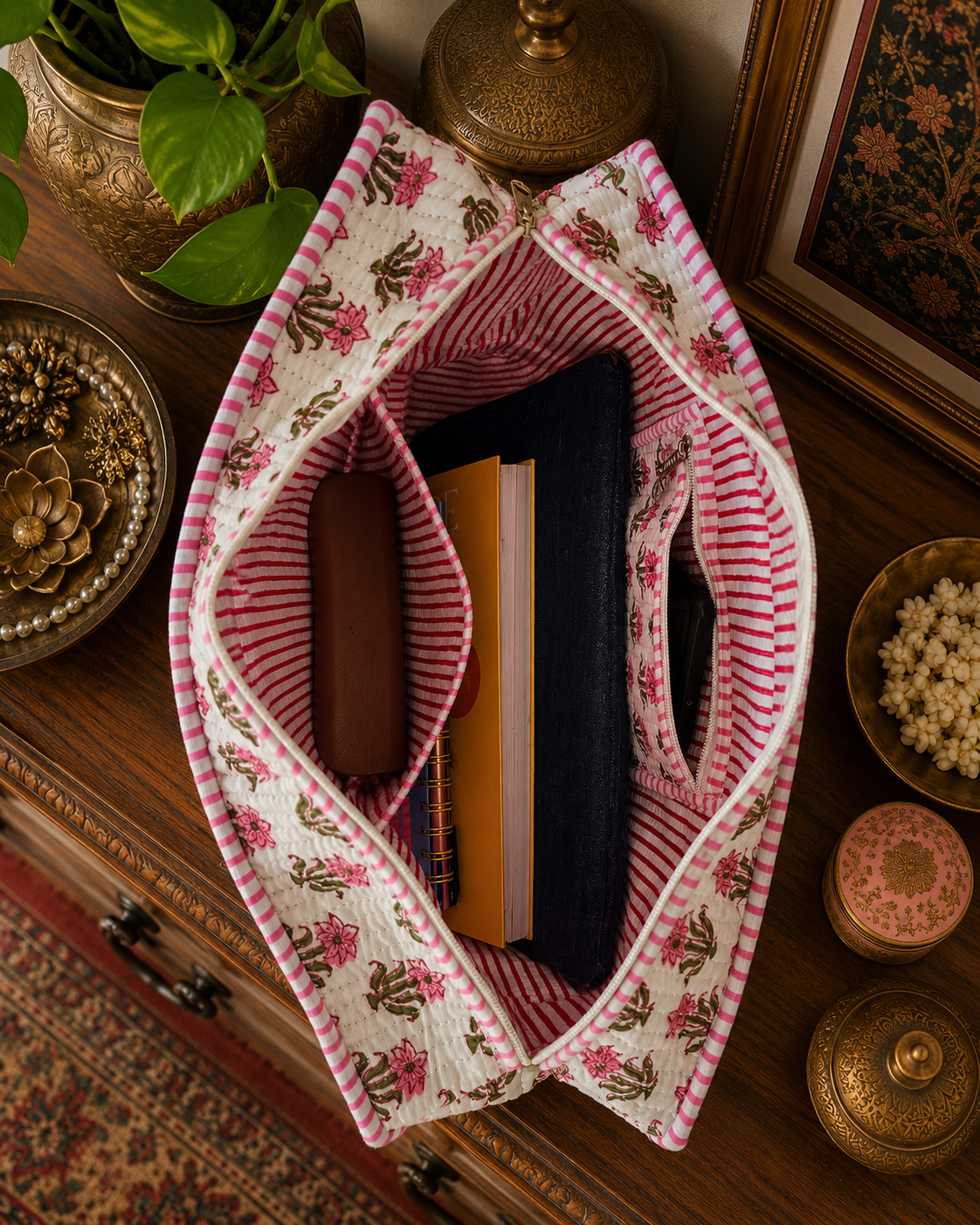 A decorative fabric quilted tote with floral patterns on a wooden surface with decorative items. The tote is carrying a laptop sleeve, notepads, sunglass cover and a mobile phone. 
