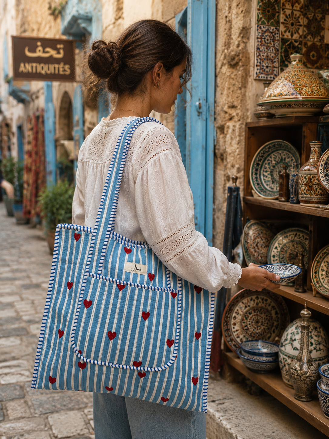 A woman is shopping in an outdoor market with a blue-striped tote bag featuring red heart patterns.