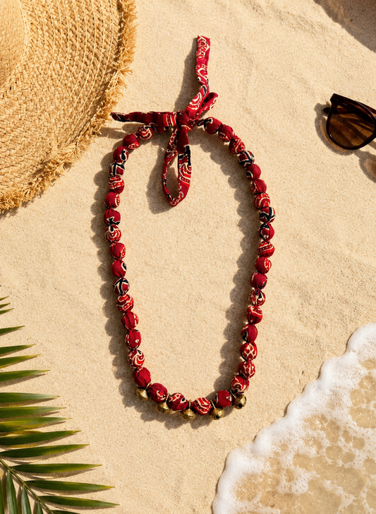 A fabric beaded Necklace with Metal bells in the centre placed on sand with other products like hat, sunglasses.