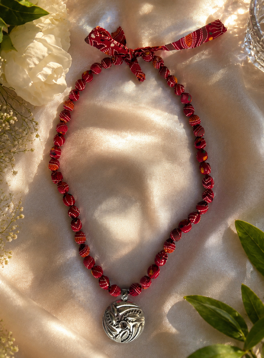 A red fabric beaded necklace with a silver pendant on a textured white surface with green leaves.