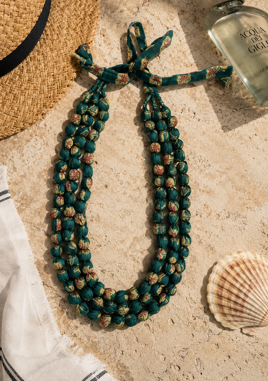 A green fabric necklace kept on a beige ceramic floor alongside shell, a hat, a perfume bottle.