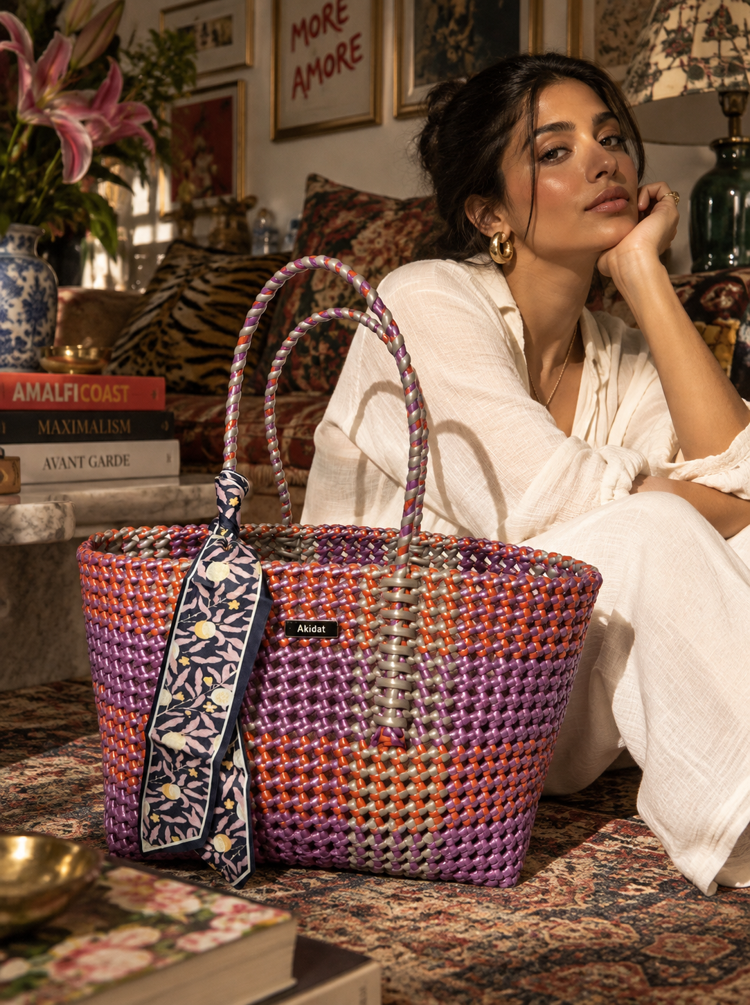 A colourful  recycled plastic woven tote bag with purple, orange, and grey stripes sits on a patterned rug in a cosy, stylishly decorated room. A woman in a white linen dress sits in the background, looking thoughtfully to the side.