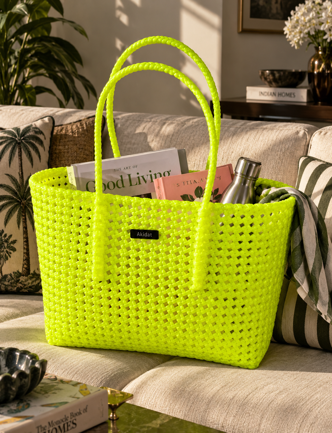 A bright neon yellow-green woven recycled plastic tote bag with rope style handles sits on a beige sofa in a styled living room. The bag contains magazines and a metal bottle and is framed by patterned pillows and home decor.