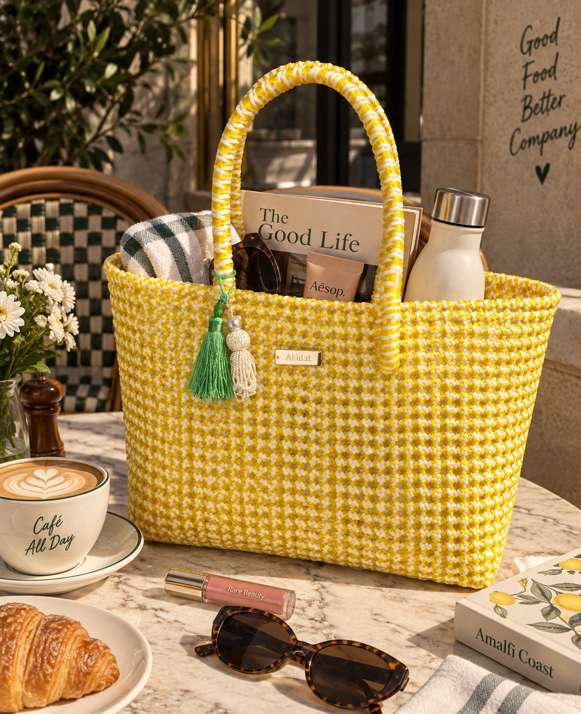 A bright yellow and white woven recycled plastic tote bag with white accents, featuring dual top handles, a green and white tassel charm, and a small white brand plaque. The bag is styled on a marble table with a coffee cup, croissant, sunglasses, and other lifestyle props.
