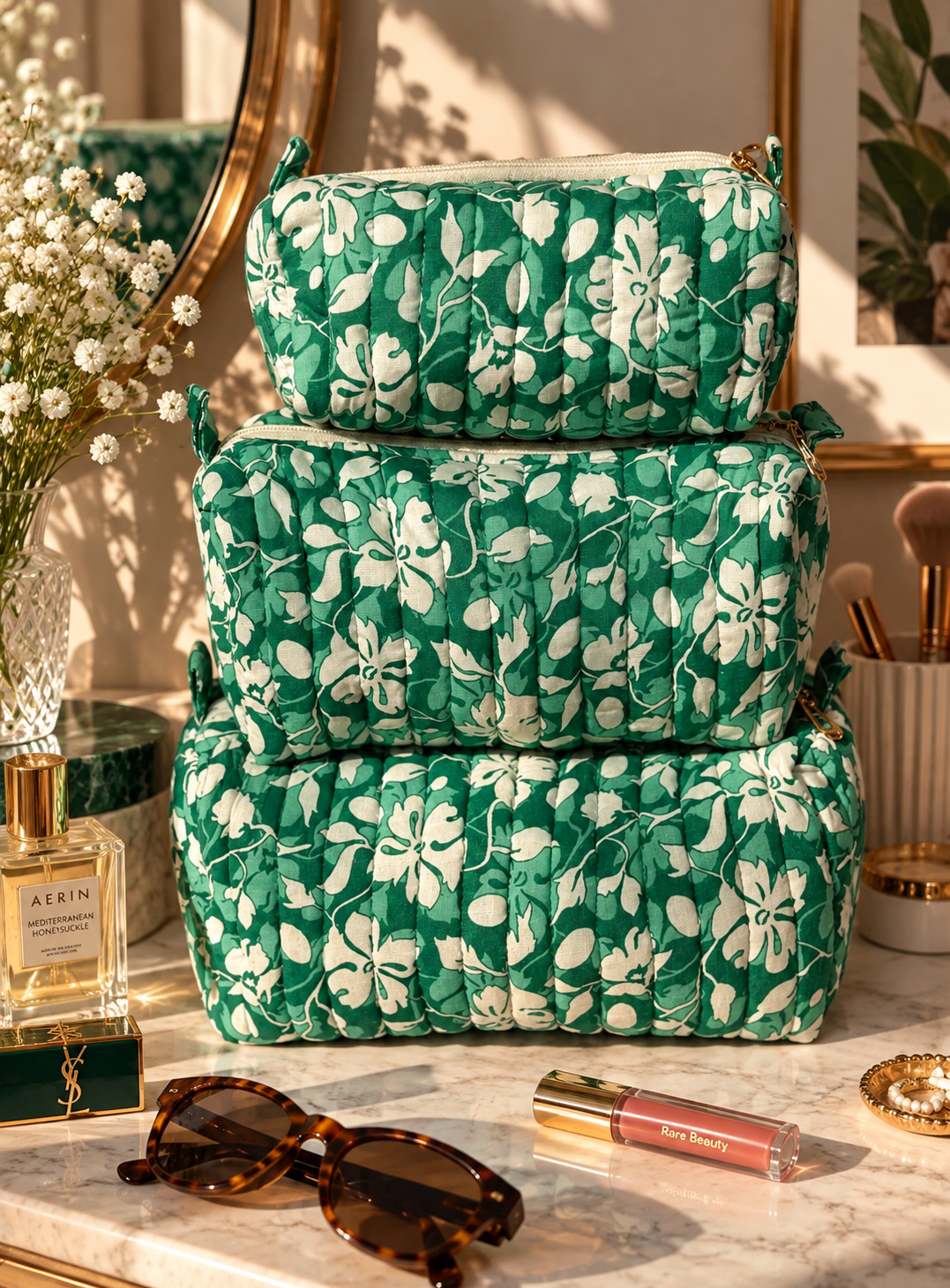Set of 3 vanity kits of different sizes resting on a marble dressing table with makeup products, brushes, and perfume, sunglasses, flowers. 