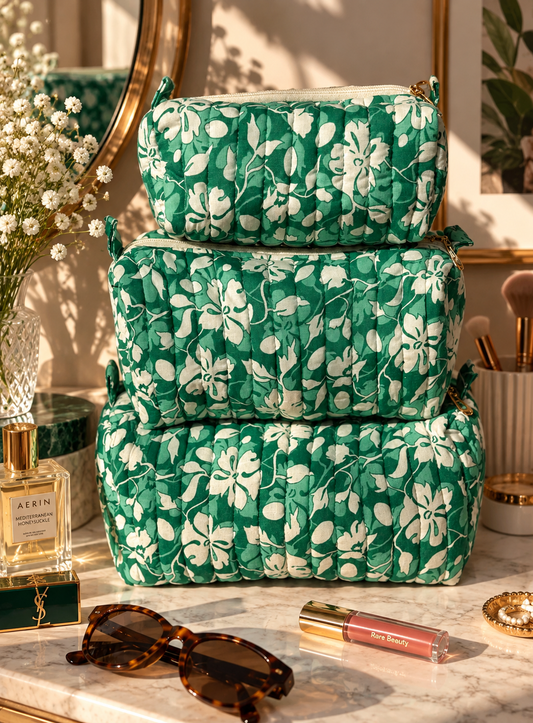 Set of 3 vanity kits of different sizes resting on a marble dressing table with makeup products, brushes, and perfume, sunglasses, flowers. 