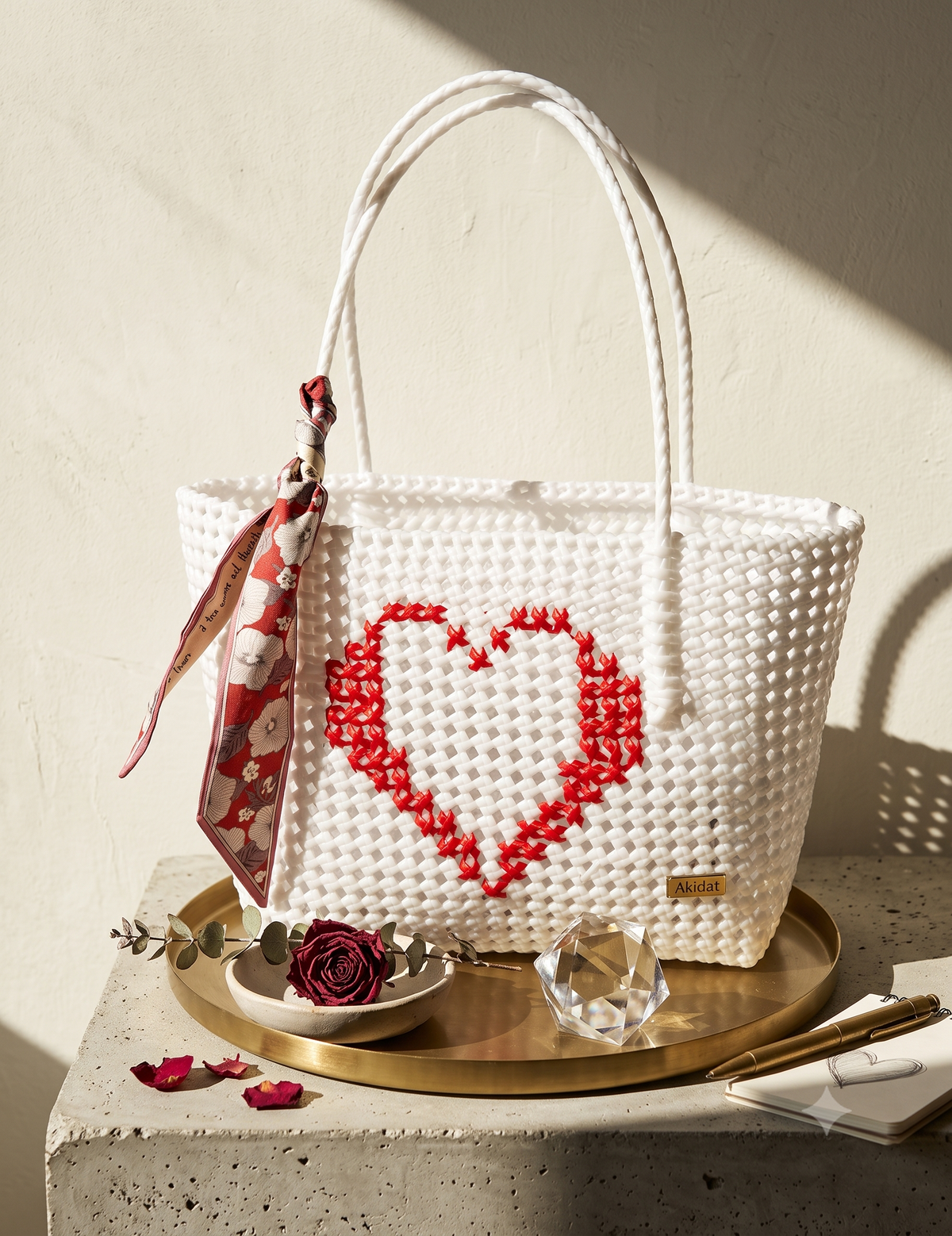 A white recycled plastic woven tote bag with a red heart design on the front, displayed on a gold tray with a patterned scarf, a rose, and a crystal on a stone surface against a textured beige wall.
