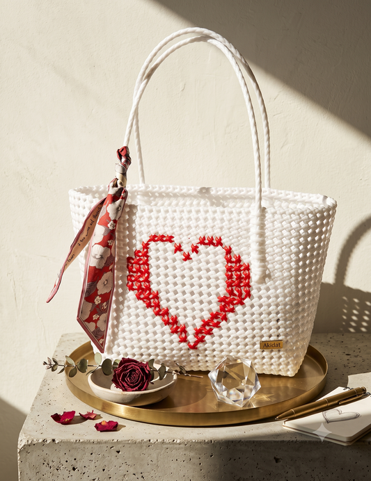 A white recycled plastic woven tote bag with a red heart design on the front, displayed on a gold tray with a patterned scarf, a rose, and a crystal on a stone surface against a textured beige wall.