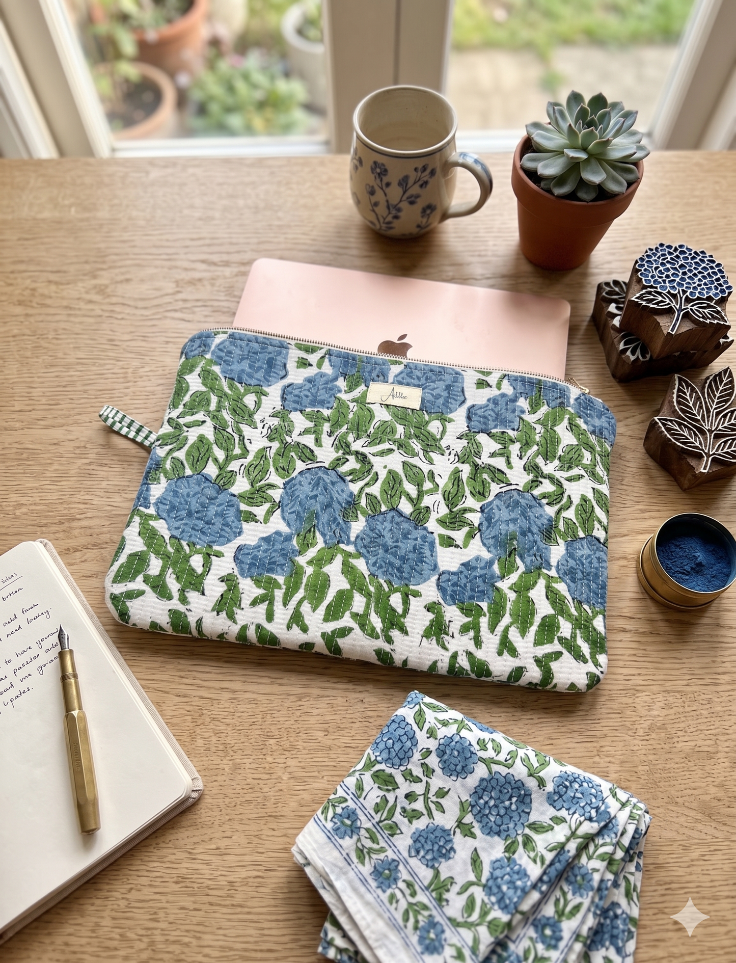 A quilted laptop sleeve with a blue-green floral pattern all over on a white base is carrying a laptop placed on a wooden table with houseplants, a coffee mug, a notebook and a pen. A similar looking cotton cloth piece placed next to it. 