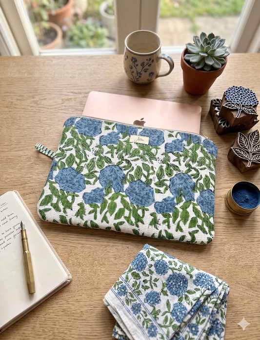 A quilted laptop sleeve with a blue-green floral pattern all over on a white base is carrying a laptop placed on a wooden table with houseplants, a coffee mug, a notebook and a pen. A similar looking cotton cloth piece placed next to it. 