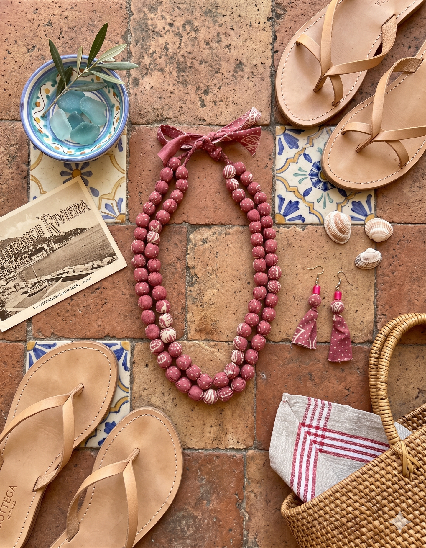 A crimson pink beaded fabric necklace with matching earrings, kept alongside with sandals, basket, and other accessories on a brick surface