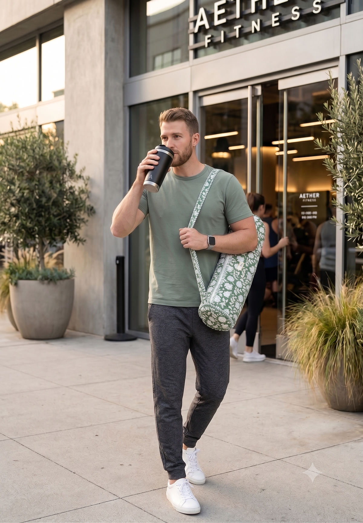 A man carrying a block print quilted yoga mat cover is coming out of a fitness center, is holding a coffee mug, and is dressed in active wear. 