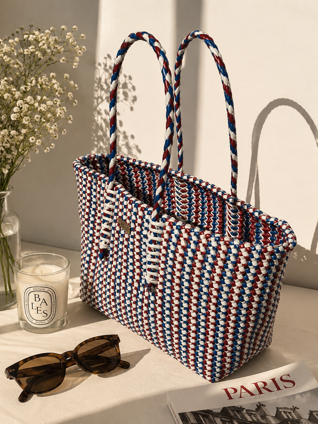 A woven recycled plastic tote bag with red, white, and blue braided handles sits on a table beside a candle, sunglasses, and a Paris magazine, with a vase of baby's breath in the background.