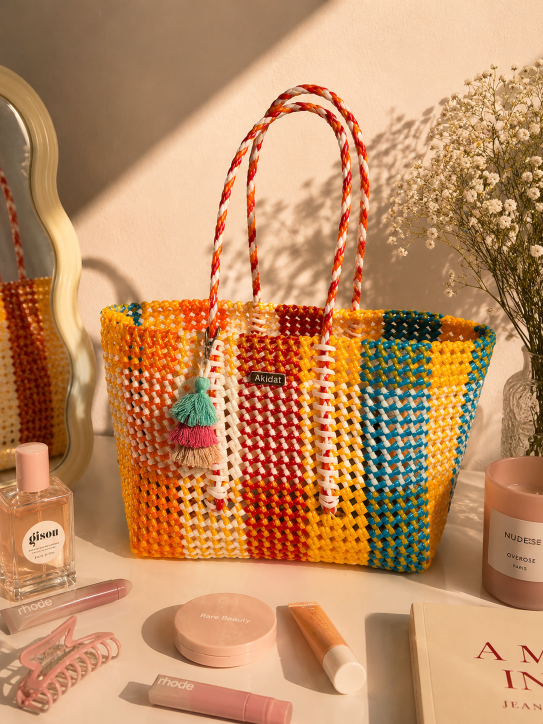A colourful recycled plastic tote bag with a charm resting on a table with other makeup products, a mirror and a flower vase. 