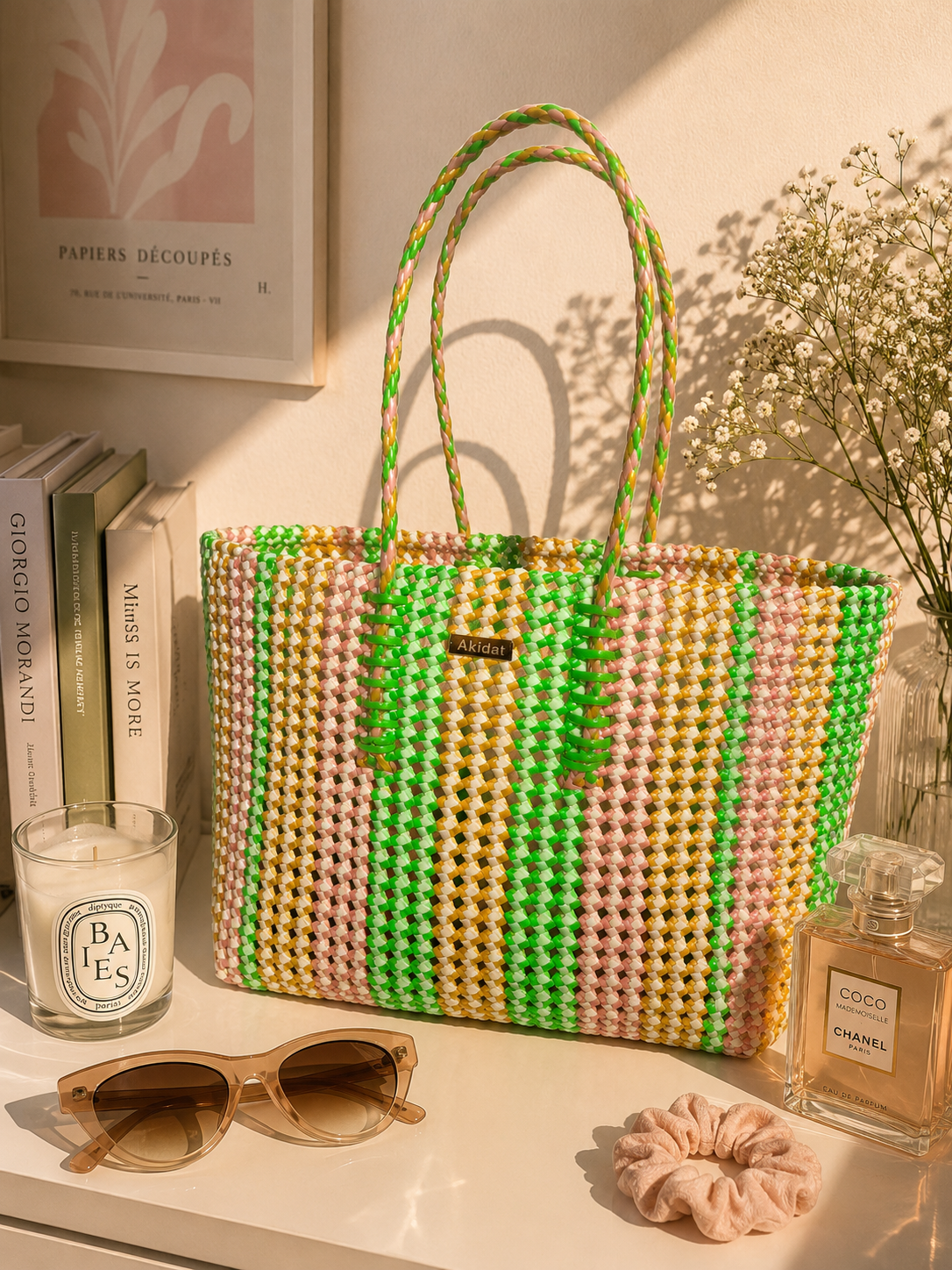 A colourful woven recycled plastic tote bag with green, orange and pink stripes sits on a light wood vanity table, with sunglasses, a perfume bottle, some books and a candle. The background shows a softly lit room with a framed art.