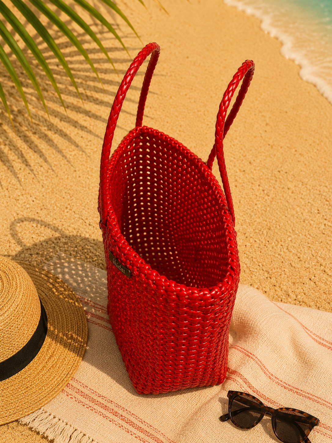 Top view of a red woven recycled plastic tote bag on a sandy beach with a straw hat and sunglasses.