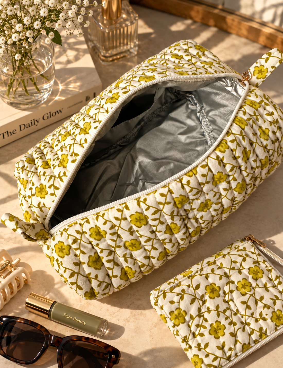 A quilted cosmetic pouch with a lemon green floral pattern, shown open with its silver-lined water-proof interior. A smaller pouch and a small rectangular item are placed beside it on a light surface, with sunglasses, a lip product, and a vase of white flowers nearby.