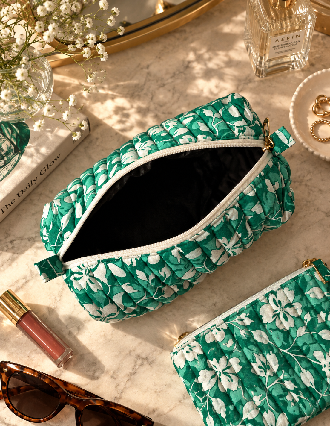 A green and white floral patterned quilted cosmetic pouch with water-proof interior and a white zipper, shown open on a marble surface with lifestyle props including sunglasses, lipstick, a perfume bottle, and flowers.