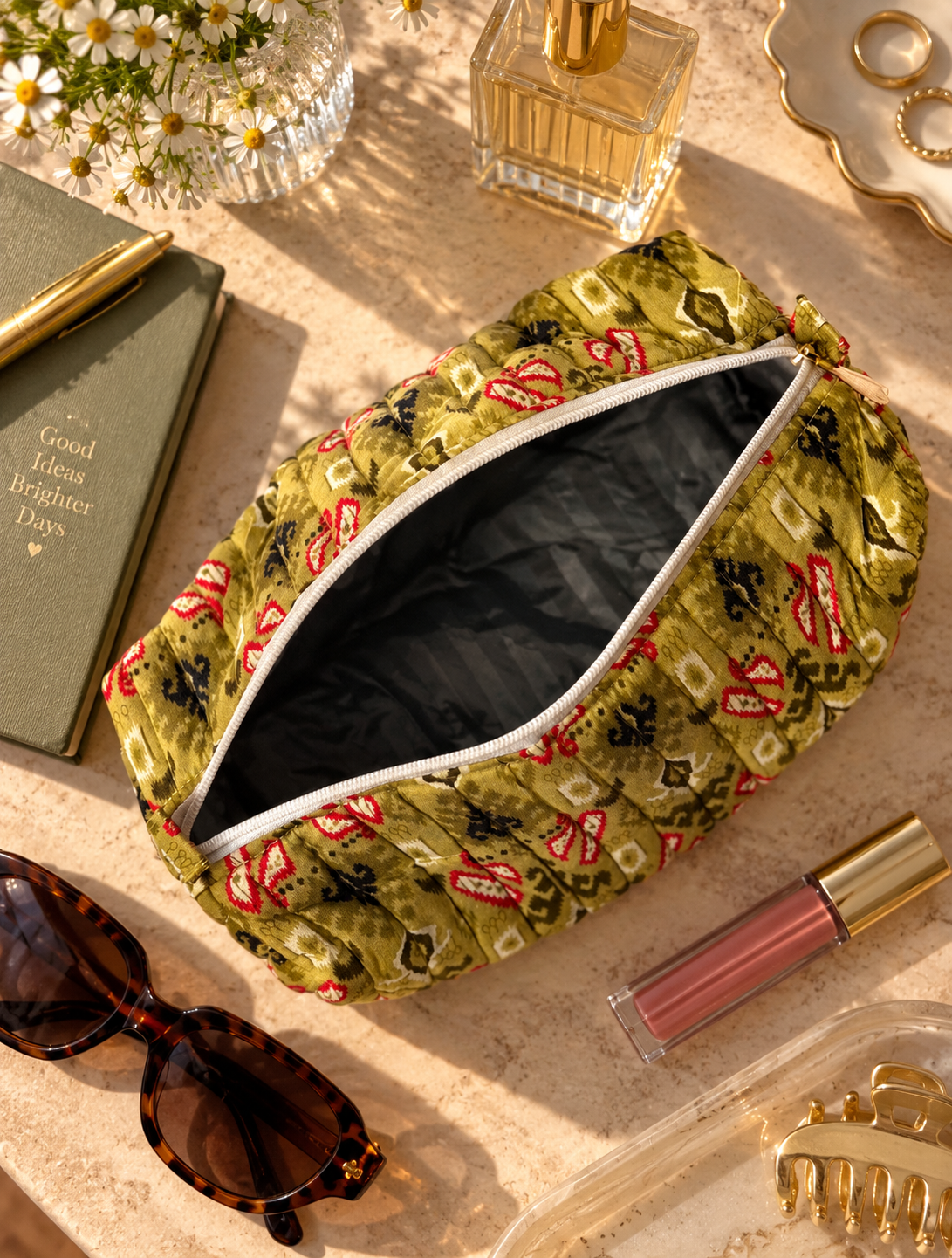 A quilted cosmetic pouch with an olive green fabric and a red abstract pattern, water-proof interior, shown open on a styled flat-lay with sunglasses, lipstick, perfume, and flowers.