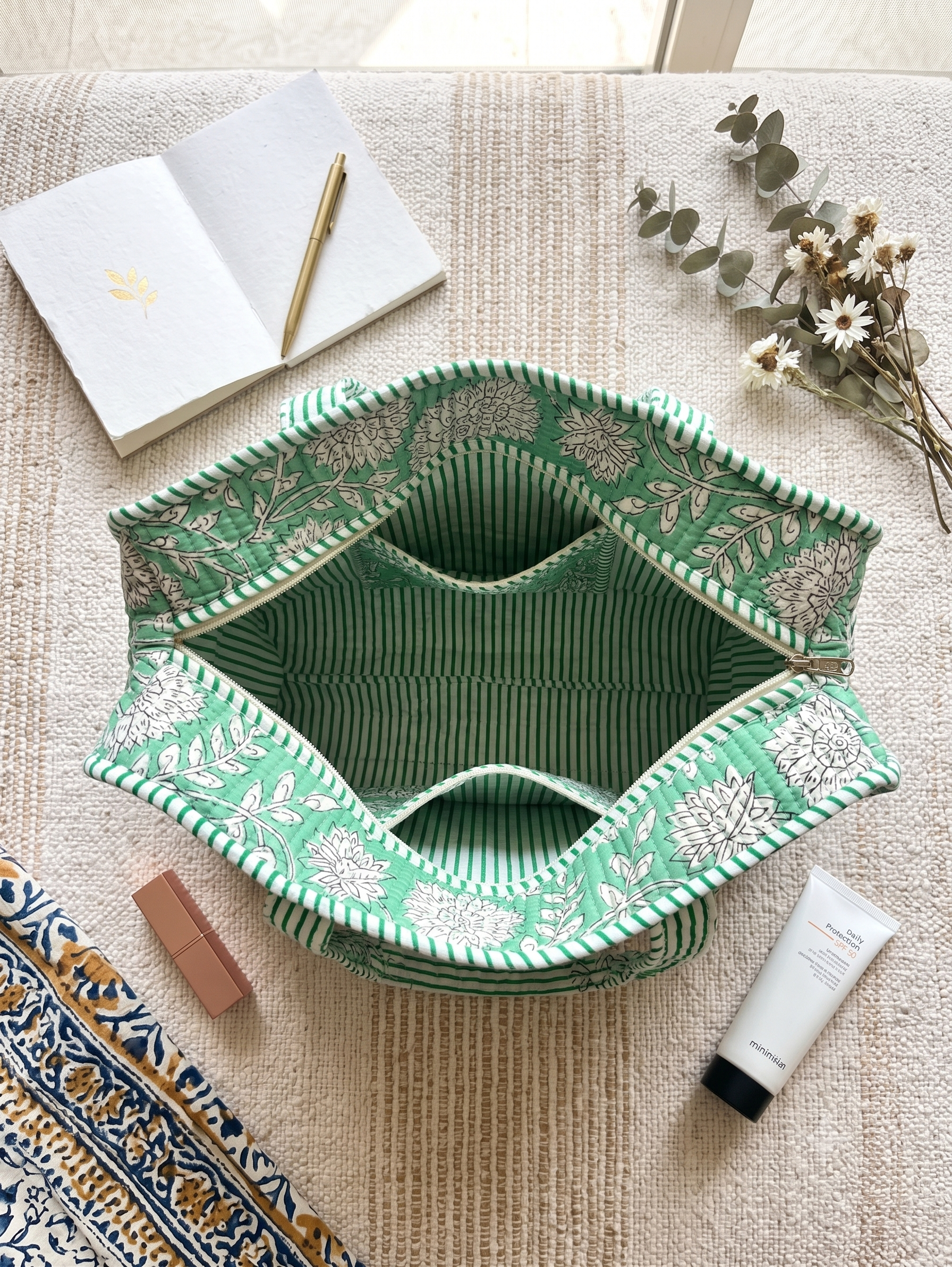 A green and white floral patterned fabric tote bag is styled in a flat lay on a beige textured blanket, surrounded by lifestyle props including a notebook, pen, dried flowers, a tube of lotion, and a lip balm. The inside of the bag is green and white stripes and showes 3 pockets. 
