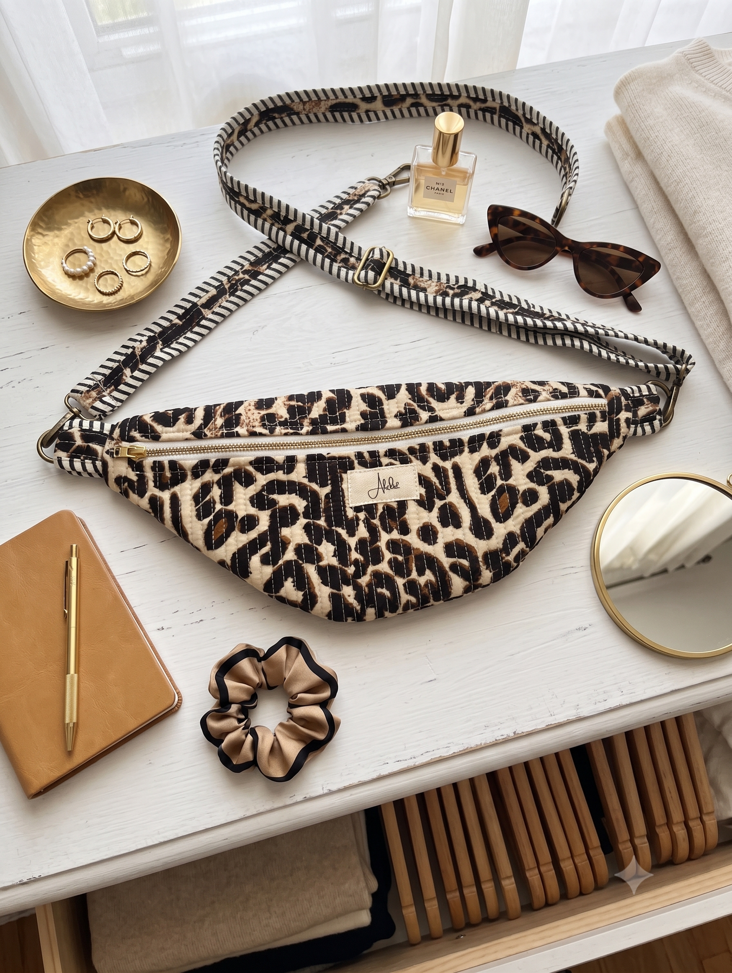 Animal print cross-body fanny pack with an adjustable strap on a white table with an accessory tray, sunglasses, a perfume bottle, a mirror, a scrunchie and a notebook. 