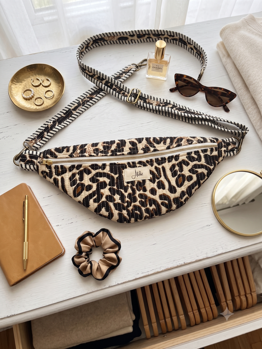 Animal print cross-body fanny pack with an adjustable strap on a white table with an accessory tray, sunglasses, a perfume bottle, a mirror, a scrunchie and a notebook. 