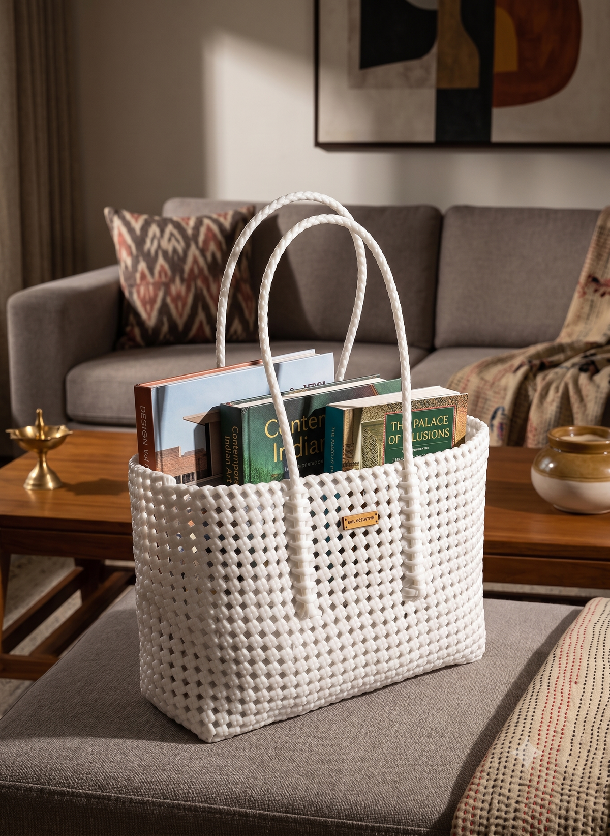 A white woven recycled plastic tote bag with rope handles sits on a grey upholstered ottoman in a living room, holding several books. A grey sofa, wooden coffee table, and framed wall art are visible in the background.