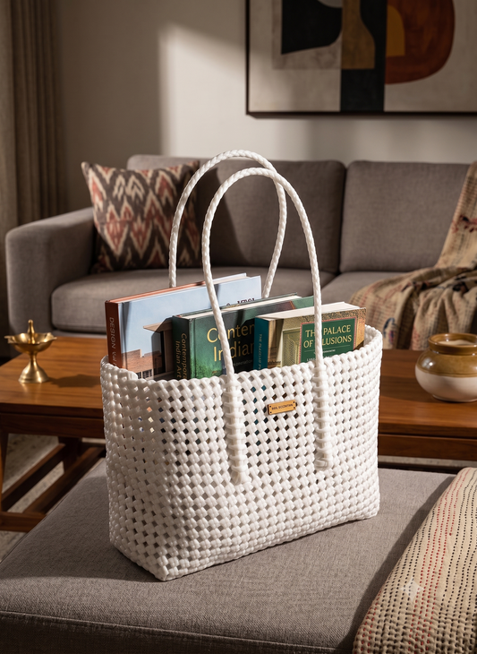 A white woven recycled plastic tote bag with rope handles sits on a grey upholstered ottoman in a living room, holding several books. A grey sofa, wooden coffee table, and framed wall art are visible in the background.
